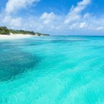Bright, clear turquoise waters of a shallow tropical sea or lagoon with white sandy beach and green vegetation under a blue sky with scattered clouds