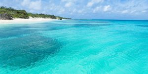 Bright, clear turquoise waters of a shallow tropical sea or lagoon with white sandy beach and green vegetation under a blue sky with scattered clouds